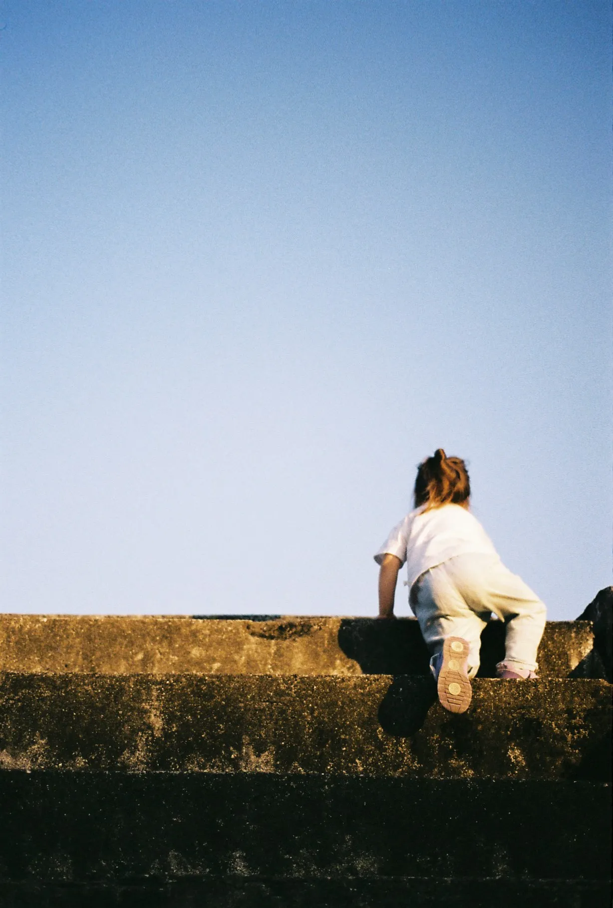 A small child seen from behind climbs over a low concrete wall under a wide blue sky, one foot lifted mid-step, with warm sunlight casting soft shadows across the textured surface.