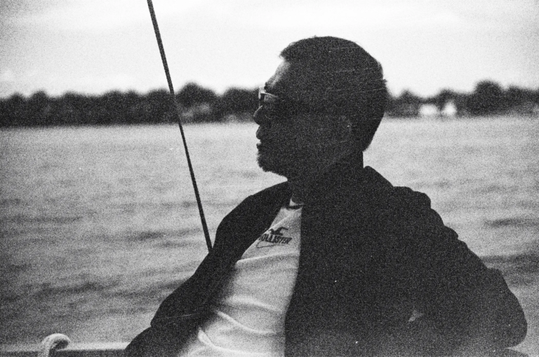 Black-and-white film photograph of a man in side profile sitting on a sailboat, wearing glasses and a jacket, holding a line or rope, with water and a distant shoreline blurred behind him. The image has visible grain and a rough texture.