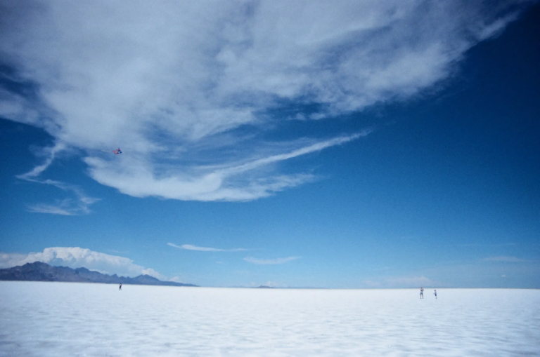 A vast white salt flat stretching to the horizon under a deep blue sky with sweeping clouds. Tiny human figures stand far away on the right, and a small kite flies in the sky, emphasizing the scale and openness of the landscape.