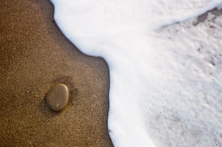 Close-up photograph of wet sand meeting white sea foam on a beach, with a small smooth pebble resting near the edge where the water touches the shore.