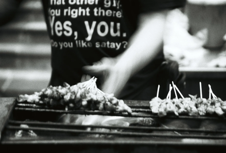 close-up of satay skewers grilling over charcoal with a vendor’s hands in the background, evening street food scene at Lau Pa Sat, film photography by Percy Lin 2016