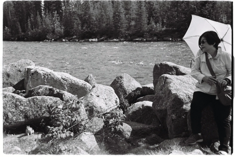 A black-and-white film photograph of a woman sitting on large rocks beside a flowing river, holding a white umbrella. A small squirrel stands on the rocks nearby, with trees and forest in the background.