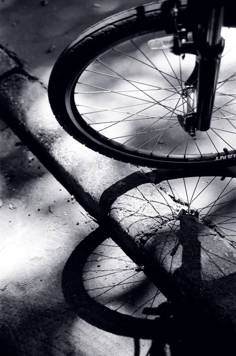 Front bicycle wheel casting a strong shadow on cracked concrete ground in black and white, film photography by Percy Lin 2014