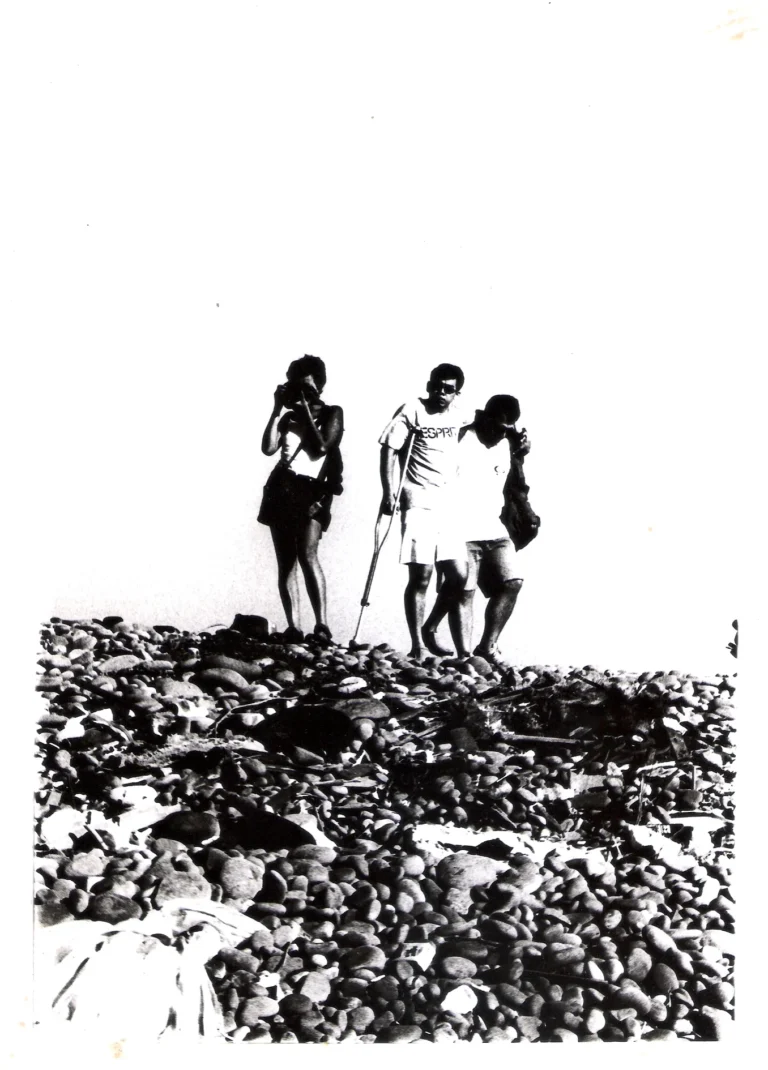 Black and white film photograph of three friends walking on a rocky shore, one using crutches while being supported by a partner — film by Scot Andersen 2000