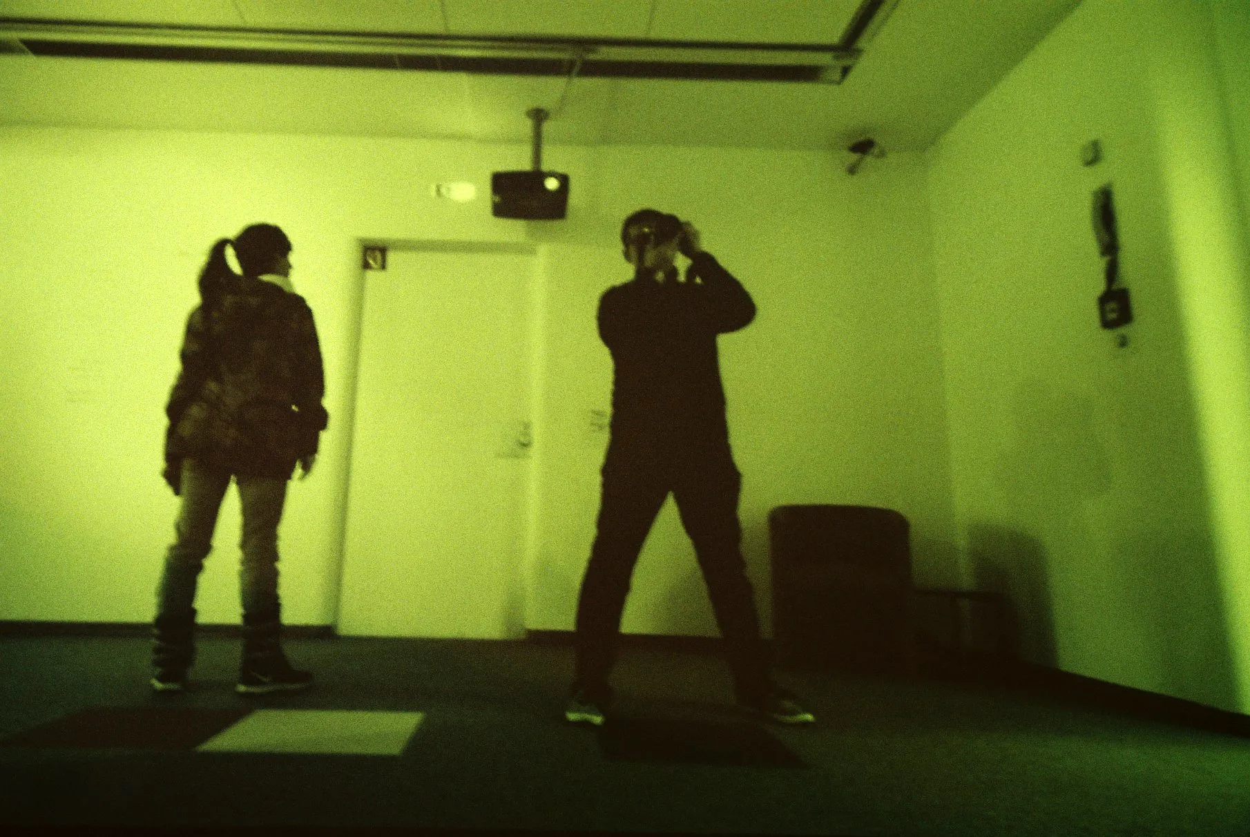 Two people standing inside a projection room with green-tinted light, one raising a camera as if observing the act of seeing — film by Percy Lin 2016