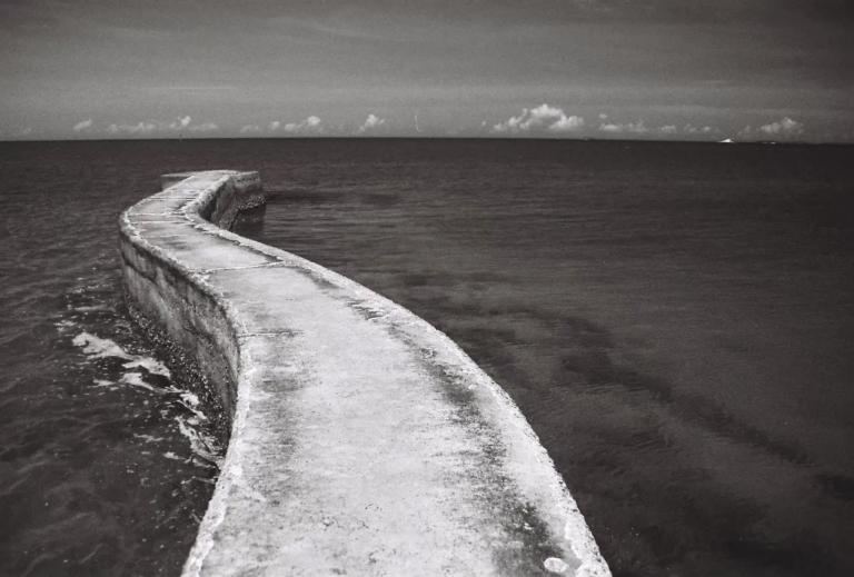 Black and white film photograph of the winding Paradise Road seawall extending into the ocean in Penghu, symbolizing the unseen foundations beneath visible choices. Film Photography by Percy Lin, 2021.